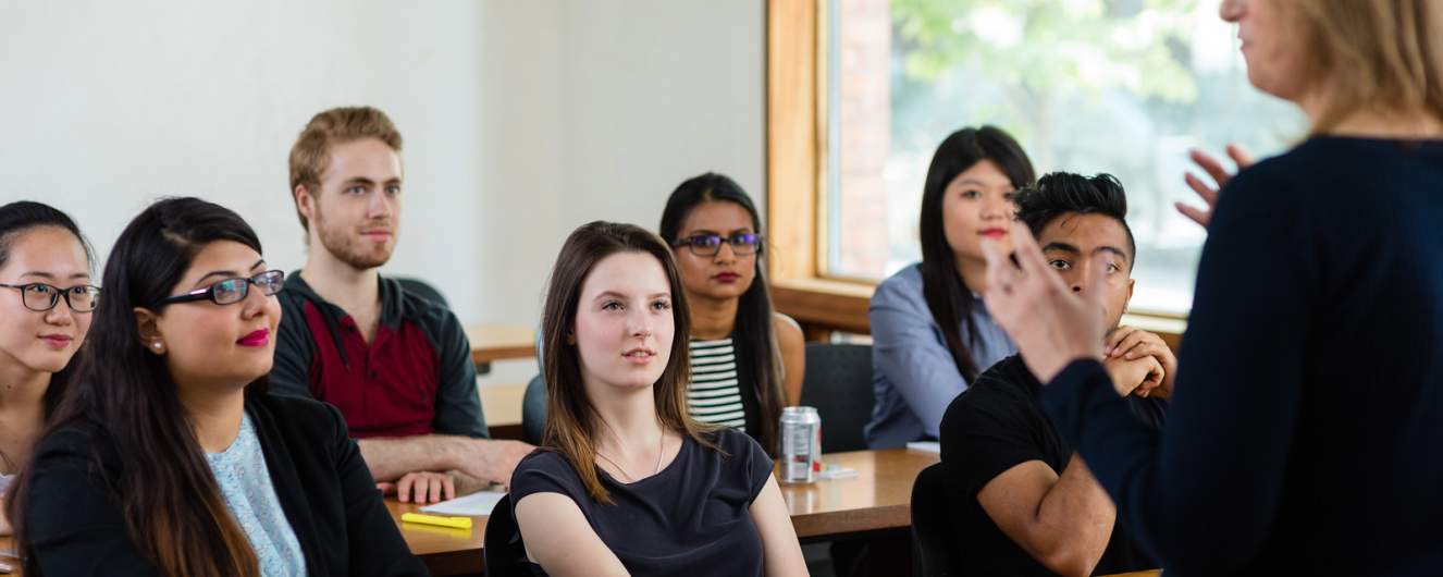 People sitting at desks, watching a speaker who is standing at the front of the room