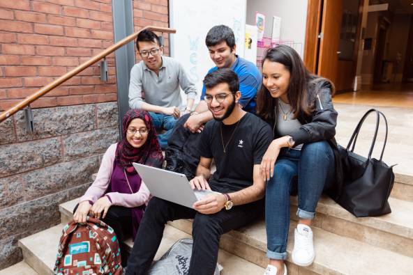 Five students sitting inside Woodsworth College looking at a laptop together and smiling